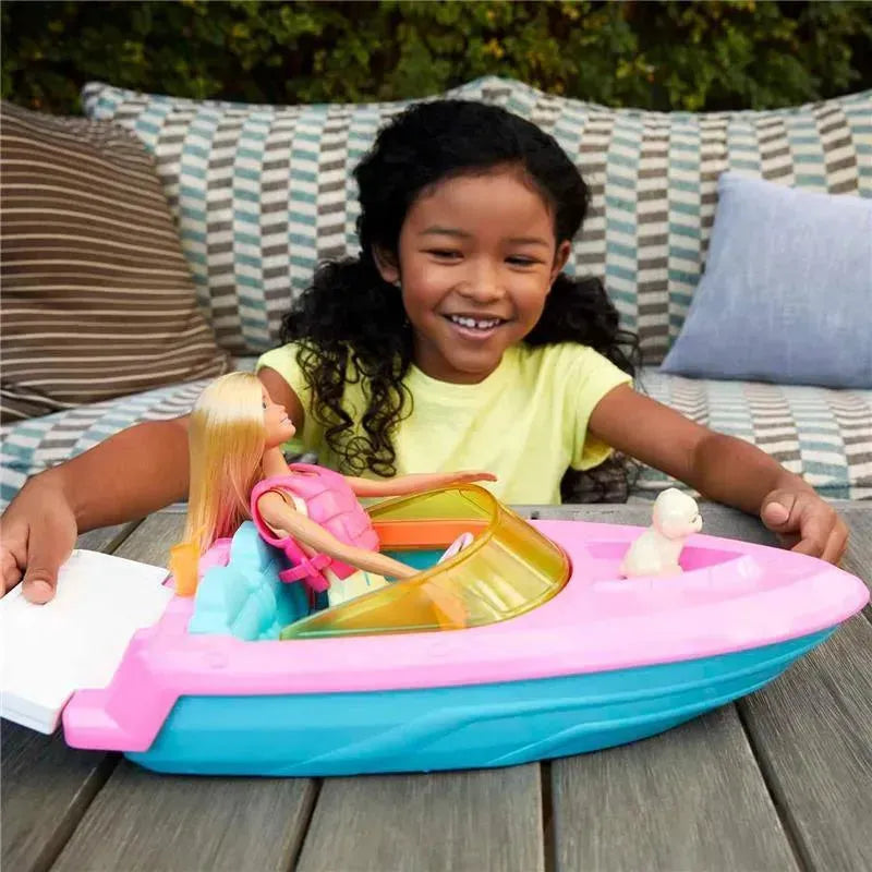 A girl with curly hair smiles while playing with a pink and blue Barbie boat that includes a doll and a small animal figure.