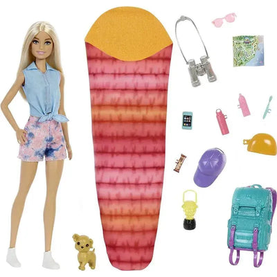 Barbie doll in a casual outfit beside a colorful sleeping bag and camping accessories including binoculars, a backpack, and a toy dog.