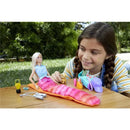 Barbie doll in a mermaid outfit sits on a table while a girl interacts with her and a small dog, surrounded by camping accessories.
