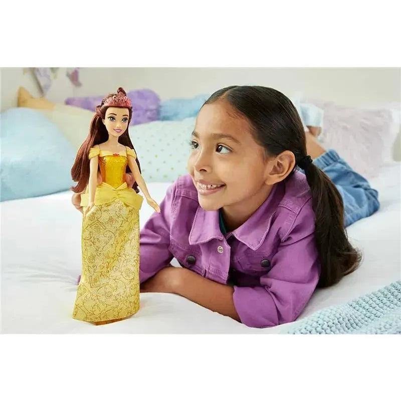 Disney Princess Belle Fashion Doll stands next to a smiling girl in a purple shirt, seated on a bed.