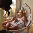Maxi-Cosi Cassia Smart Baby Swing in Truffle with an infant engaged by a caregiver and mobile toys overhead.