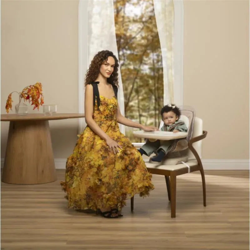 A woman in a floral dress sits beside a child in the Maxi-Cosi Kiskadee 360 Rotating Highchair, positioned in a bright, modern room.