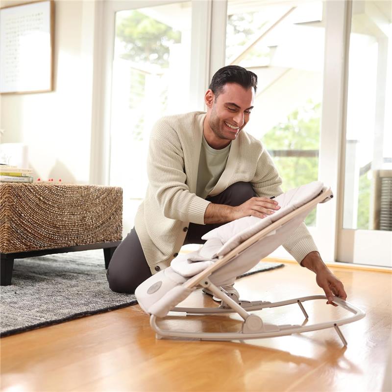 A man adjusts the Maxi-Cosi Kori 2-in-1 Baby Bouncer Seat for Infants, featuring a beige fabric and sturdy base in a bright room.