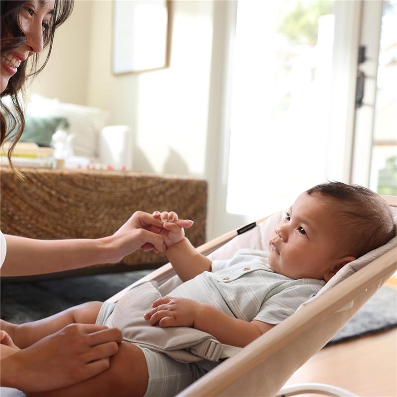 A smiling adult holds the hand of an infant seated in a beige Maxi-Cosi Kori 2-in-1 Baby Bouncer, in a bright room.