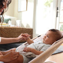 A smiling adult holds the hand of an infant seated in a beige Maxi-Cosi Kori 2-in-1 Baby Bouncer, in a bright room.