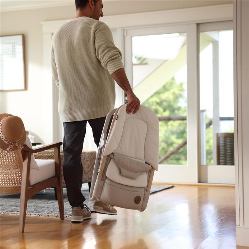 A man holds a folded Maxi-Cosi Kori 2-in-1 baby bouncer seat in a living room, featuring a light brown color.