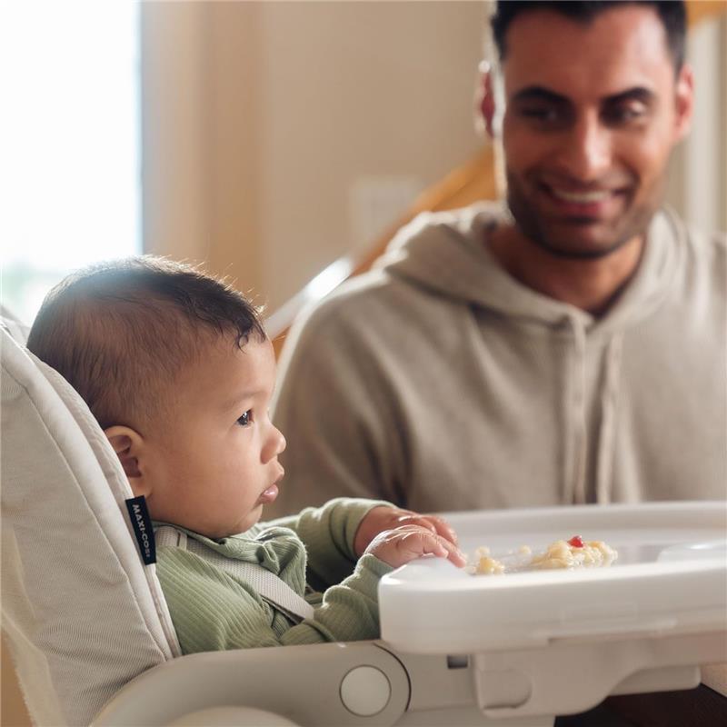 Baby in a Maxi-Cosi Minla 6-in-1 high chair, wearing a green outfit, gazes at a smiling adult while holding food.