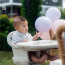 Baby sitting in a Maxi-Cosi Moa 8-in-1 high chair, looking at a floating bubble, with pastel balloons in the background.