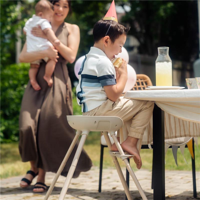 A child wearing a party hat sits on a Maxi-Cosi Moa high chair, enjoying food at an outdoor table while a woman holds a baby.