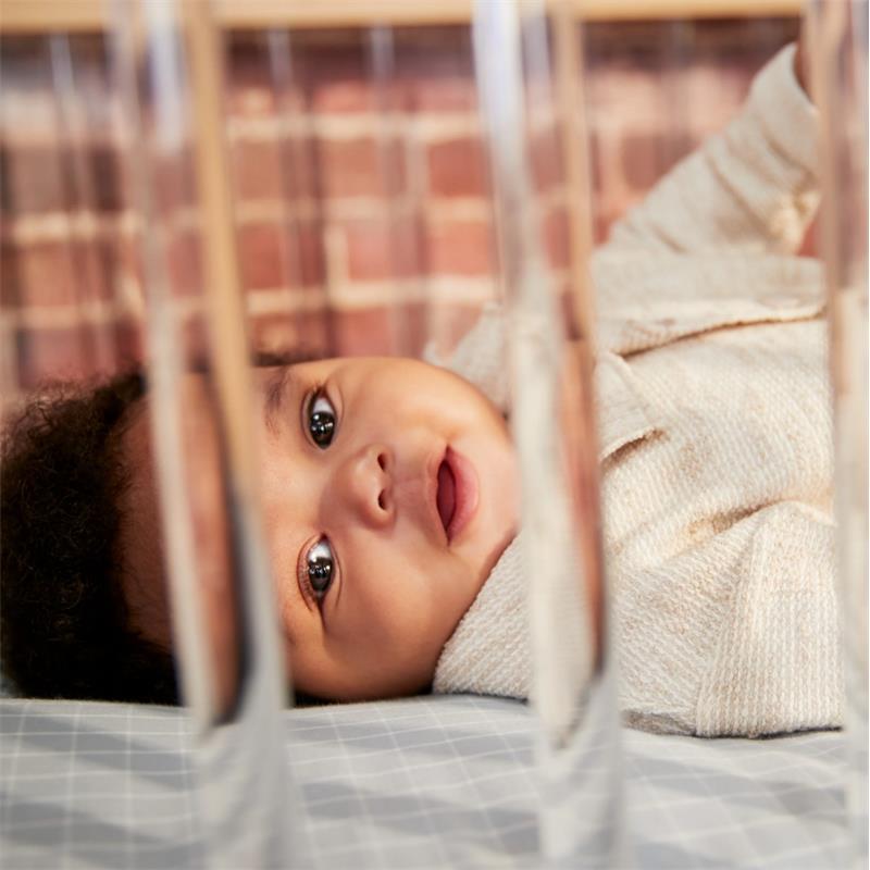 A smiling baby lays in a crib with a soft blanket, viewed through transparent crib slats, against a brick wall background.