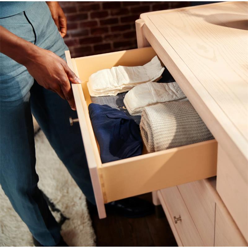 Hand reaching into a drawer of the Maxi-Cosi Nursery Brisa 6-Drawer Dresser, revealing folded clothes and diapers.