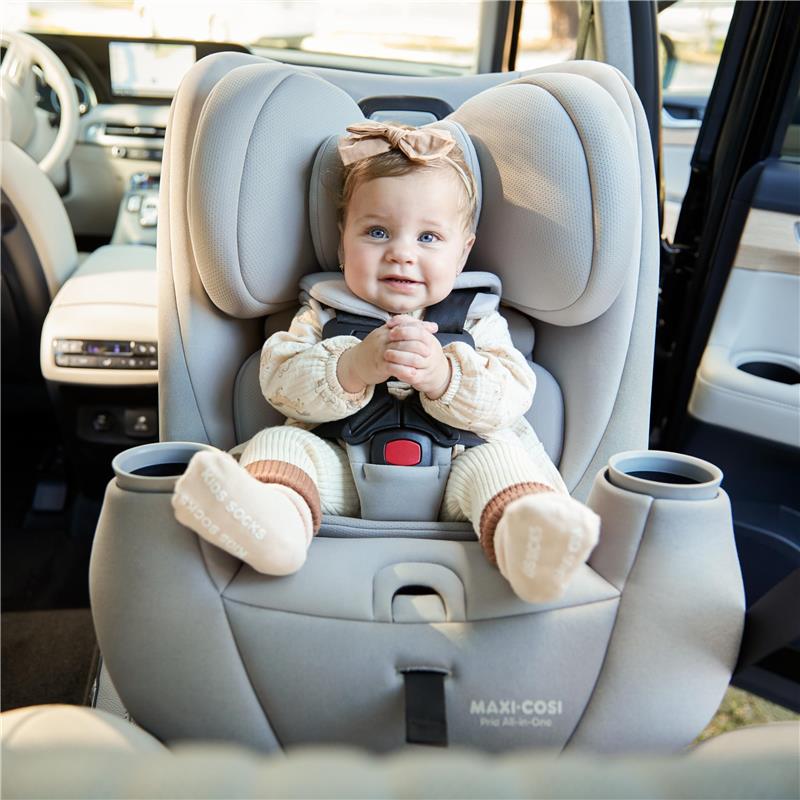 A smiling baby sits in a Maxi-Cosi Pria All-in-One Convertible Car Seat in Blackened Pearl, with a cozy interior visible.