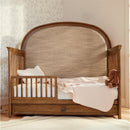 Maxi Cosi Quiet Luxury 6-in-1 Convertible Crib in natural wood finish, featuring a toddler bed configuration with a pink blanket and plush toy.
