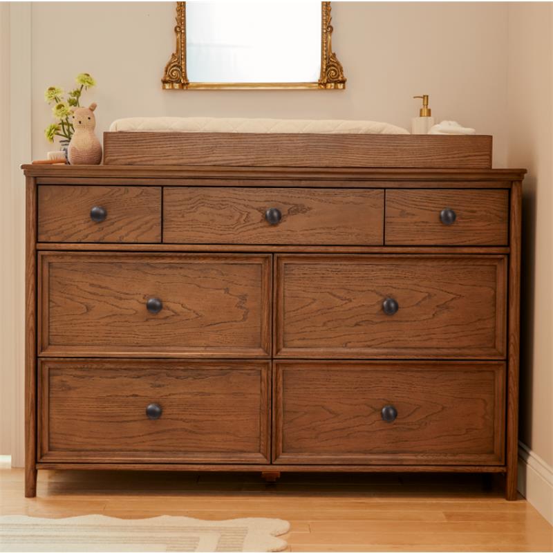Maxi Cosi Quiet Luxury 7-Drawer Dresser in a warm wood finish with round black knobs, topped with a changing pad and decorative items.