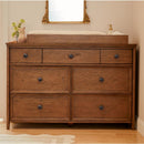 Maxi Cosi Quiet Luxury 7-Drawer Dresser in a warm wood finish with round black knobs, topped with a changing pad and decorative items.