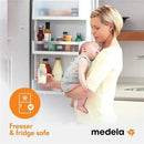 Breast milk collection and storage bottle by Medela, held by a woman with a baby in her arms, standing by an open refrigerator. Freezer and fridge safe label visible.