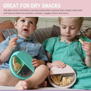 Two children sitting on a couch, each holding a Melii Spin Snack Container, one pink and one teal, filled with snacks.