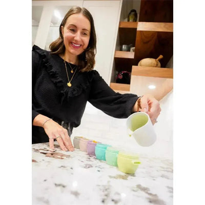 Woman pouring food into colorful Melii baby food storage containers on a kitchen countertop. Containers are 4 oz. in size.