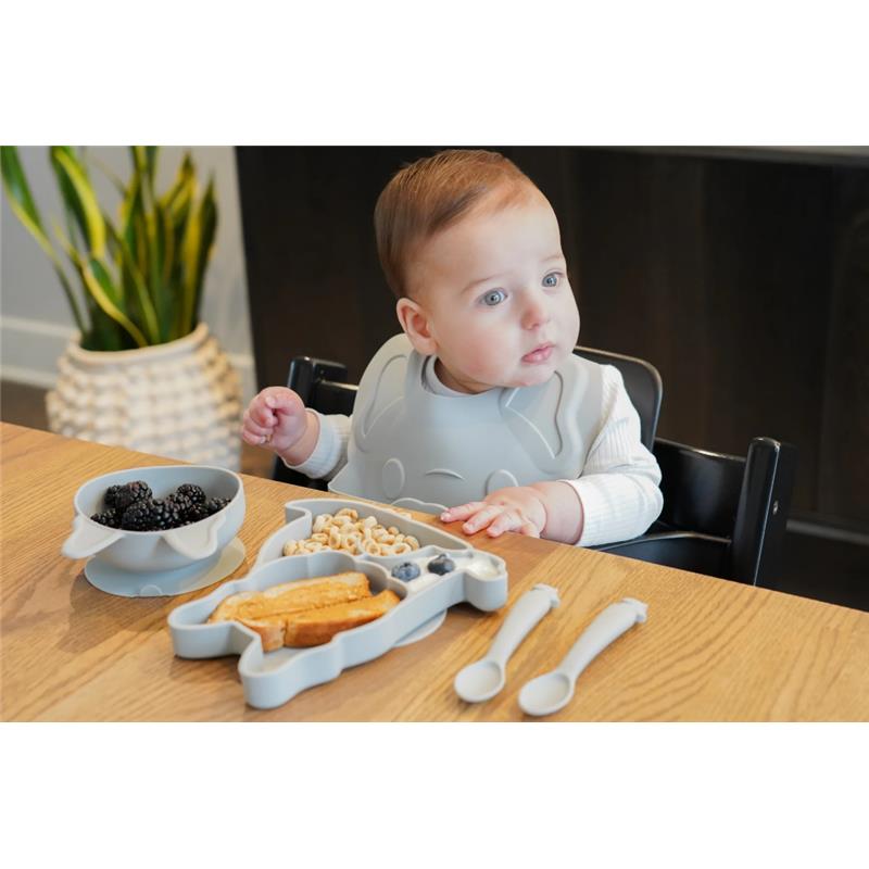 A baby in a high chair wearing a gray bib sits at a wooden table, with a suction plate shaped like a bulldog, a bowl of blackberries, and two spoons.