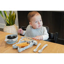 A baby in a high chair wearing a gray bib sits at a wooden table, with a suction plate shaped like a bulldog, a bowl of blackberries, and two spoons.