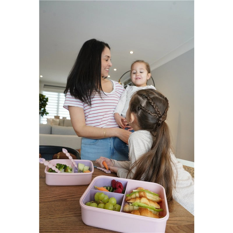 Melii Bento Box in pink and purple with removable divider, filled with fruits and sandwiches, on a table with a mother and two children.