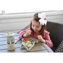 A child with a white bow in her hair uses a fork to eat from a divided silicone suction plate shaped like a bulldog, filled with sushi, carrots, and cucumber, beside a water bottle on a table.