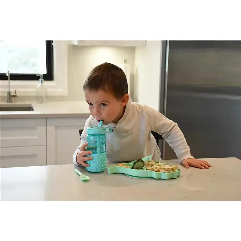 Child sitting at a table drinking from a blue silicone cup while eating from a green silicone suction plate shaped like a dinosaur.