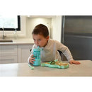 Child sitting at a table drinking from a blue silicone cup while eating from a green silicone suction plate shaped like a dinosaur.