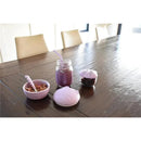 Silicone cat bowl, lid, and utensils in lavender color displayed on a wooden table with food and a drink beside them.