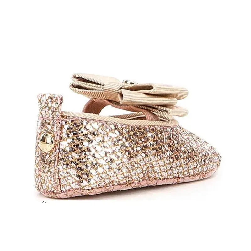 Rose gold ballet crib shoe by Michael Kors with a sparkly finish and a decorative bow on top.