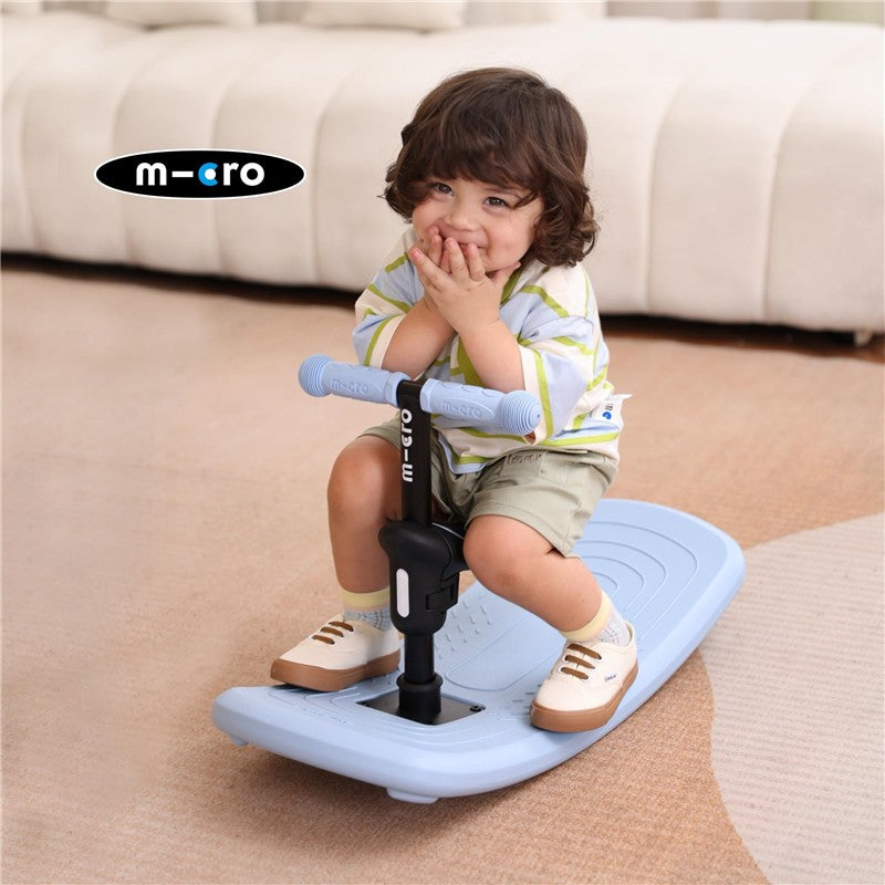 Child sitting on blue Micro Kickboard Mini Rock & Go LED scooter, smiling with hands on cheeks, in a cozy indoor setting.