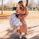 Woman in a pink dress holds a Milk Snob Disney 5-In-1 cover with a Fairytale Friends design, in a park setting.