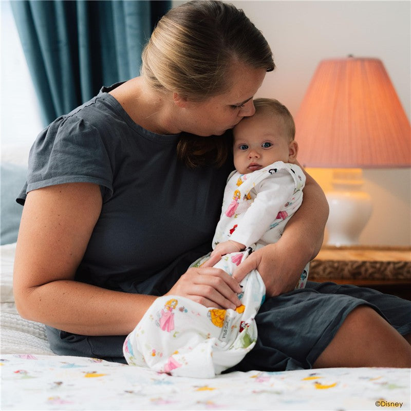 A mother holds her baby wrapped in a Milk Snob Disney Princess Sleep Bag, featuring colorful princess designs.