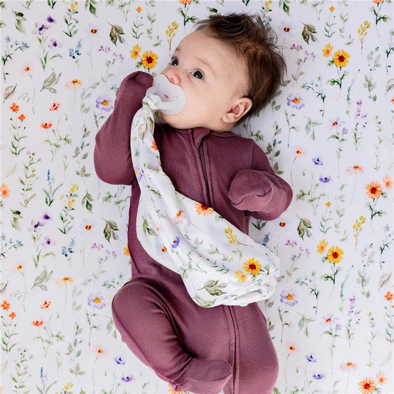 Baby lying on a floral-patterned blanket, holding a Milk Snob Eloise Lovey security blanket and a pacifier.