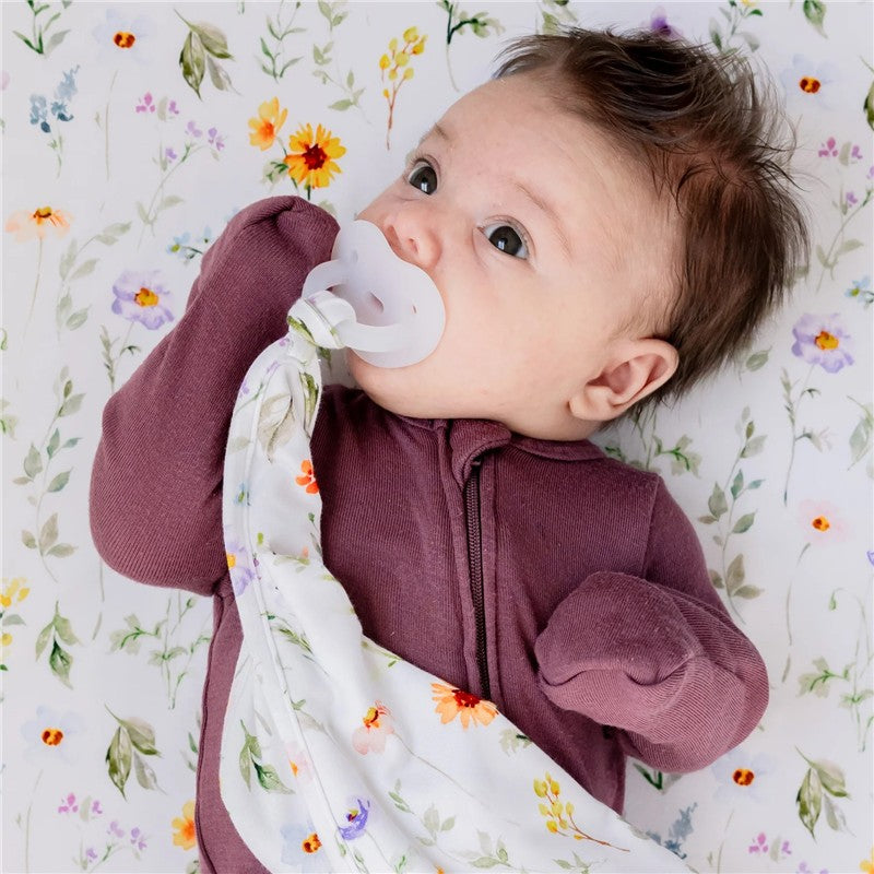 Baby lying on a floral-patterned blanket, holding a Milk Snob Eloise Lovey security blanket and sucking a pacifier.