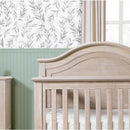 Rustic 4-in-1 Convertible Curve Top Crib in Sandbar finish, showcased in a nursery with leaf-patterned wallpaper and green paneling.