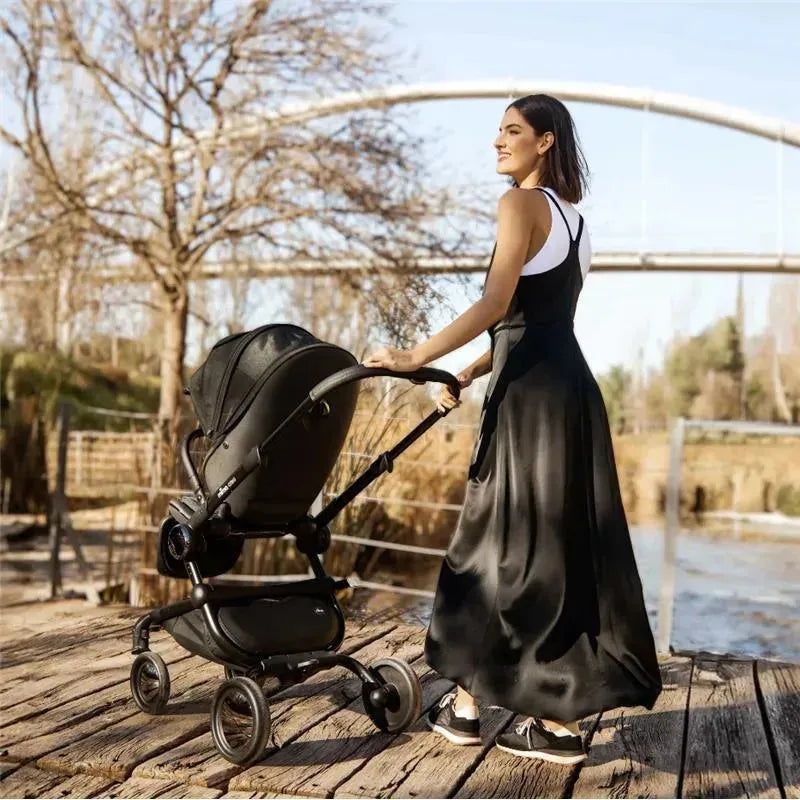 Mima Creo Single Stroller in black is being pushed by a woman in a long black dress, set against a natural backdrop.