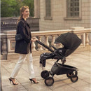A woman in a black coat and white pants strolls with a black Mima Creo Single Stroller, featuring a large canopy and stylish design.