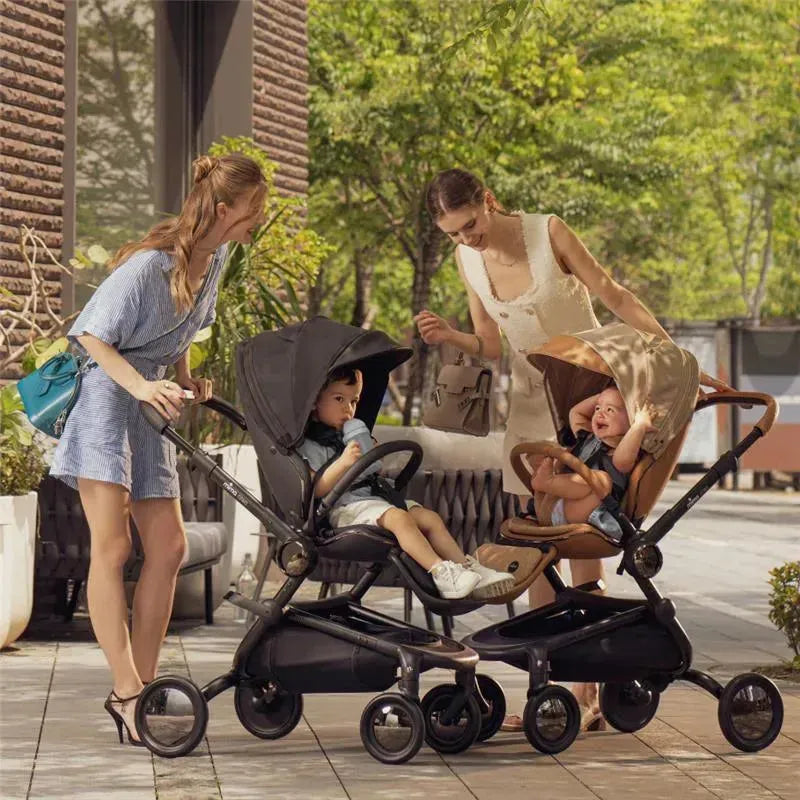 Two women interact with children in Mima Creo single strollers, one black and one brown, in an outdoor setting with greenery.