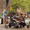 Two women interact with children in Mima Creo single strollers, one black and one brown, in an outdoor setting with greenery.