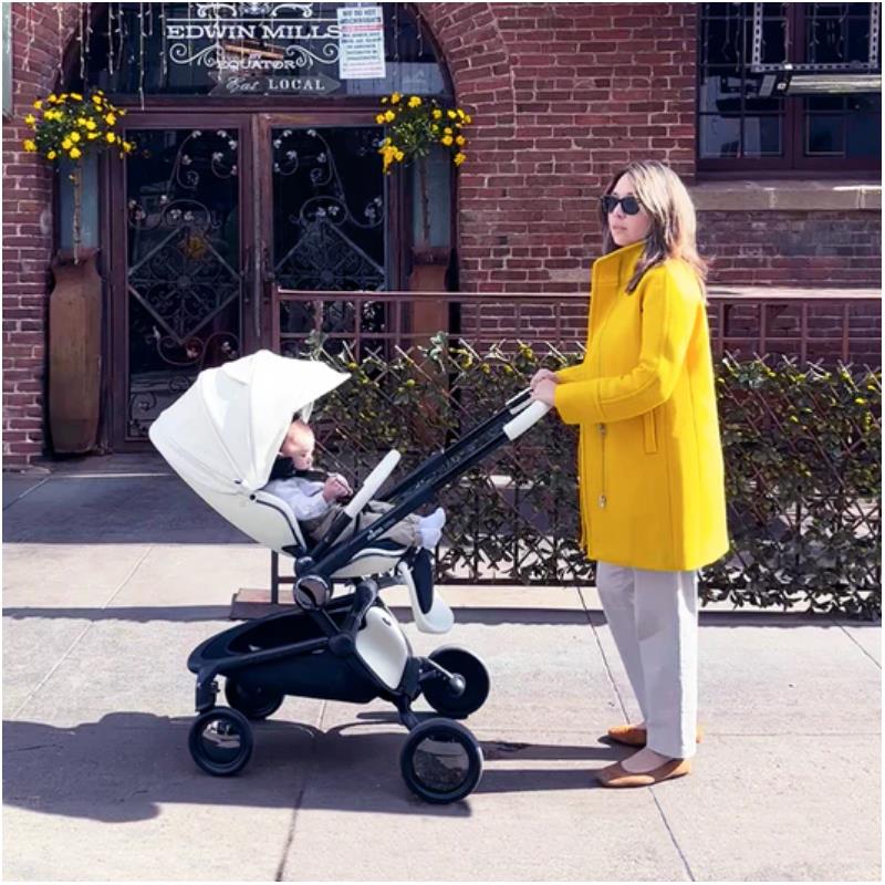 A woman in a yellow coat pushes a Mima Creo single stroller with a child inside, outdoors near a brick building.