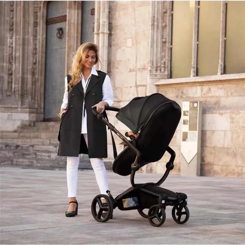 Mima Xari Max stroller in black and camel, being pushed by a woman in a stylish outfit on a city street.