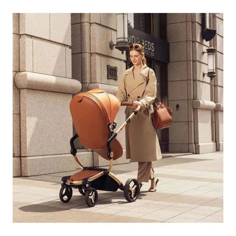 A woman in a tan trench coat pushes a Mima Xari Max stroller in black and camel while carrying a brown bag.