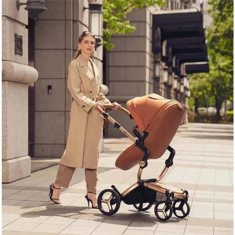 Woman in a beige trench coat walks with a Mima Xari Max stroller in black and camel colors on a city sidewalk.