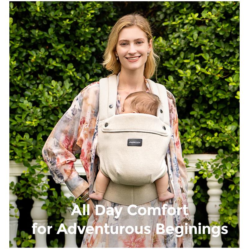 Momcozy baby carrier in light amber being worn by a smiling woman holding a baby, with greenery in the background.