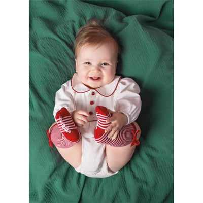 Baby wearing a white outfit with red trim, smiling while sitting on a green background, featuring red and striped socks.