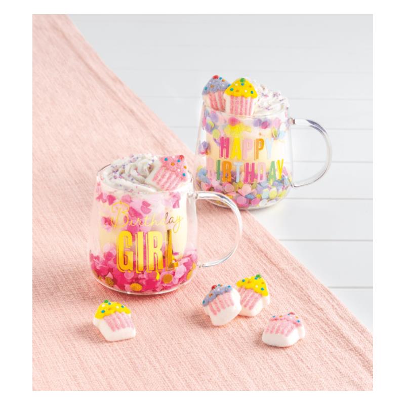Glass mugs with birthday-themed designs filled with colorful marshmallow toppings and candy cupcakes on a pink backdrop.