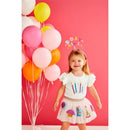 A smiling girl in a ruffled white tee adorned with colorful birthday candles, paired with a pink skirt featuring party motifs, stands among vibrant balloons.