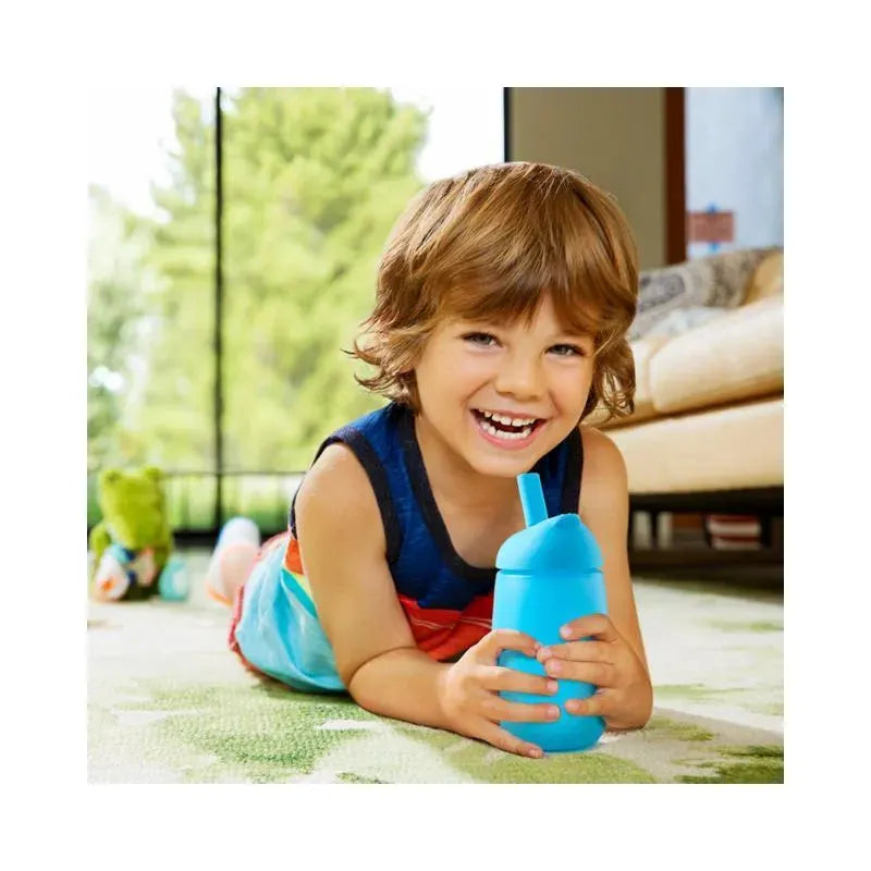 A young child smiles while lying on a colorful rug, holding a blue Munchkin 10Oz Simple Clean Straw Cup.