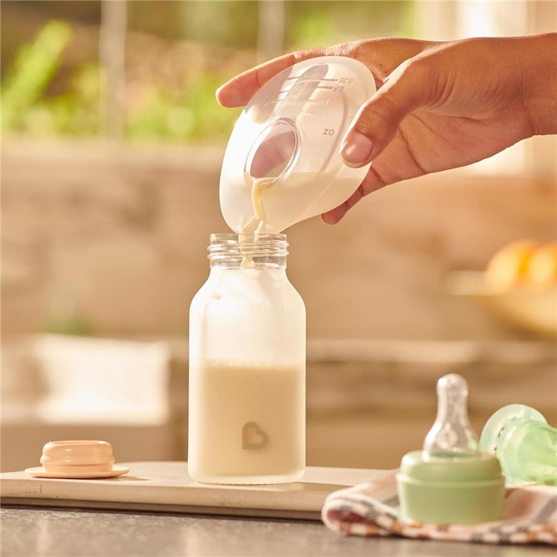 Silicone breastmilk collector cup pouring milk into a baby bottle, with a pacifier and storage container nearby.